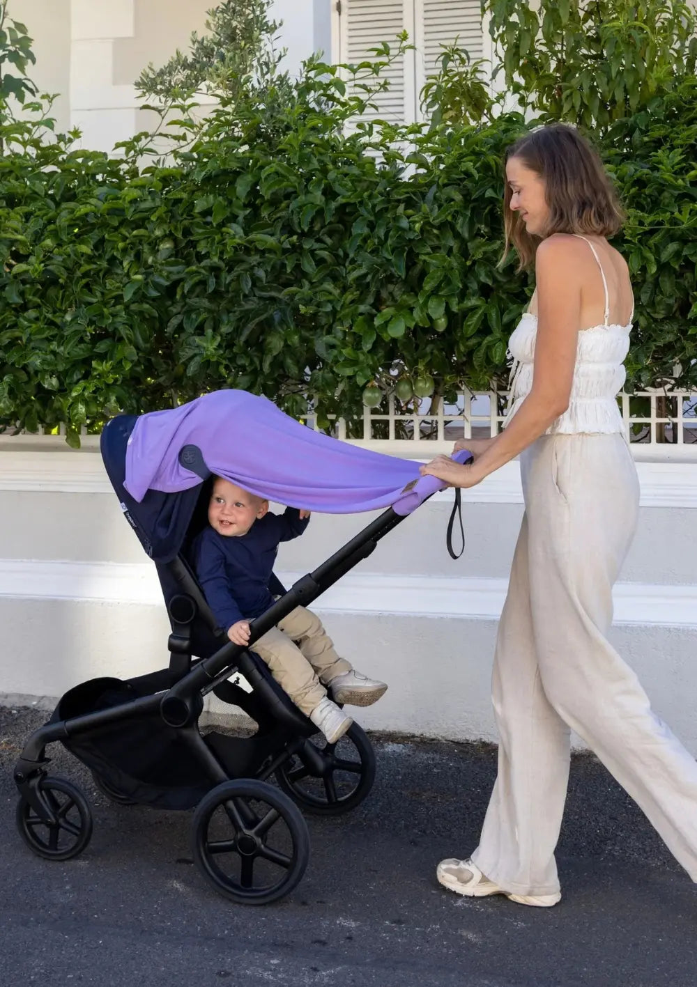 Cloby Multifunktionale UV-Decke UPF 50+ Lavender Bloom mit 2er-Set Magnet-Clips Leder Navy Blue am Kinderwagen befestigt – Frau schiebt Kinderwagen, Junge sitzt sonnengeschützt im Kinderwagen