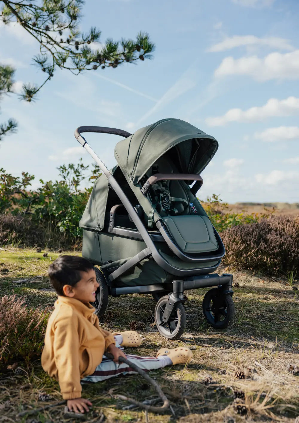 Junge sitzt auf Wiese in der Natur – im Hintegrund steht der Joolz Geschwisterkinderwagen-Set 'Geo5 Double' Forest Green