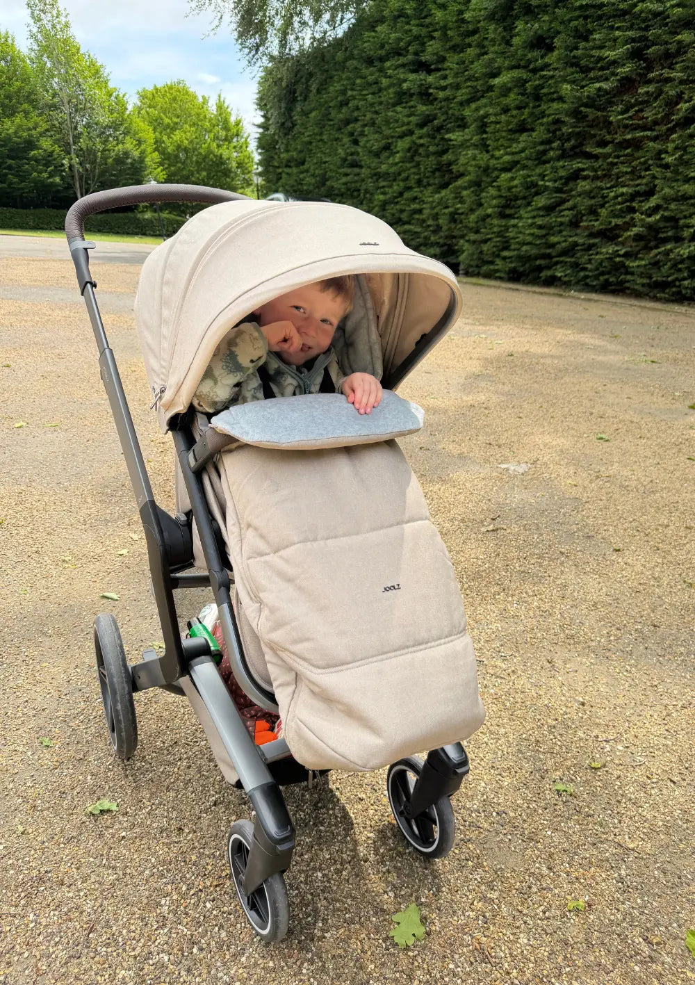 in fröhliches Kind sitzt geschützt im beigefarbenen Joolz universeller Fußsack V3 Sandy Taupe in einem passenden Kinderwagen im Freien.